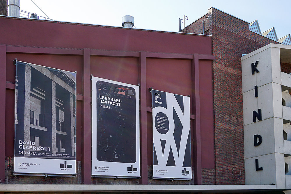 Die Ostfassade des KINDL - Zentrmu für zeitgenössische Kunst in rotem Backstein mit drei Plakaten der aktuellen Ausstellungen.